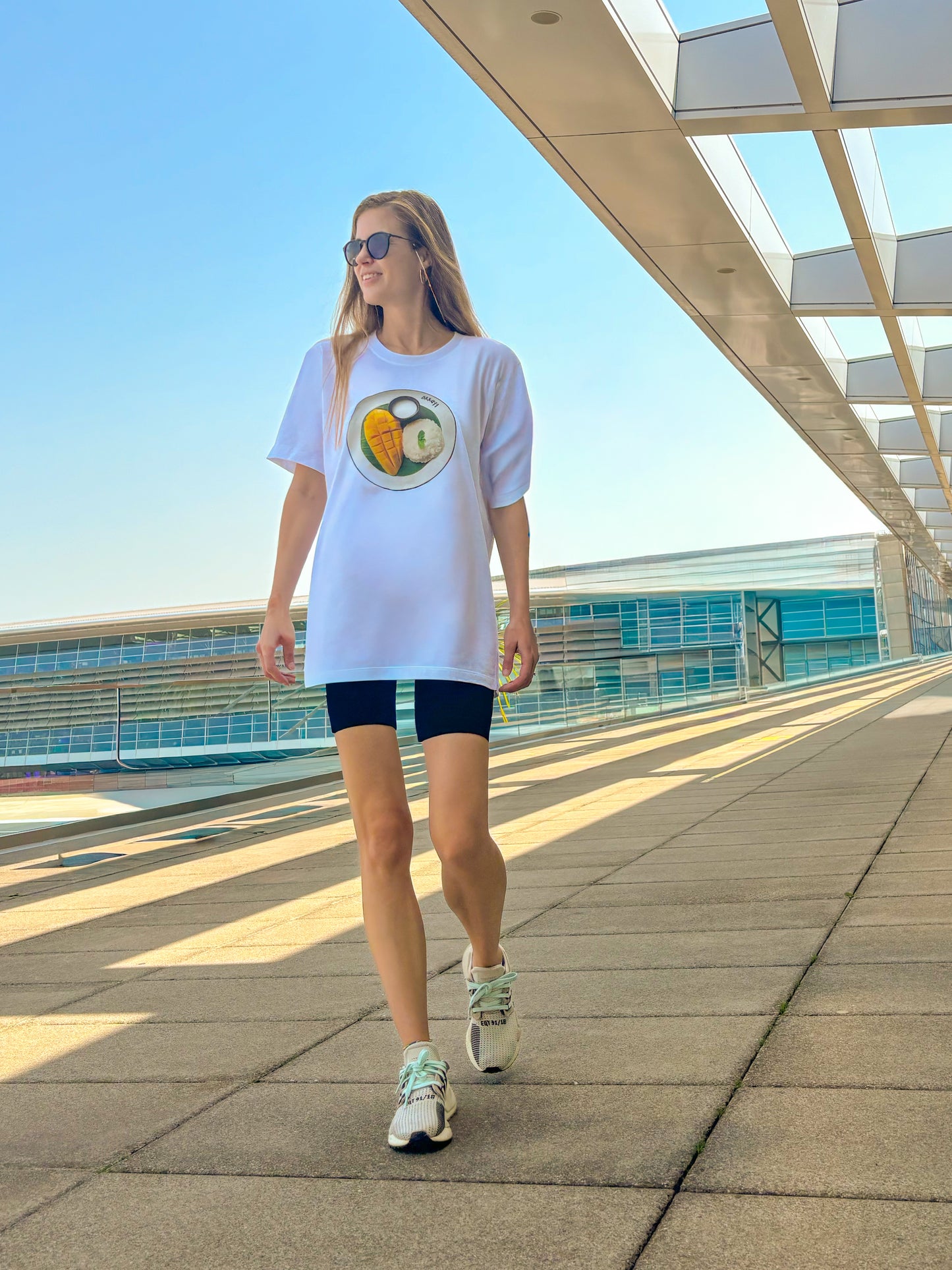 Weisses Unisex T-Shirt mit Mango Sticky Rice Food-Print mit passendem Brillenband, getragen von Frau im Sonnenlicht am Flughafen Zürich