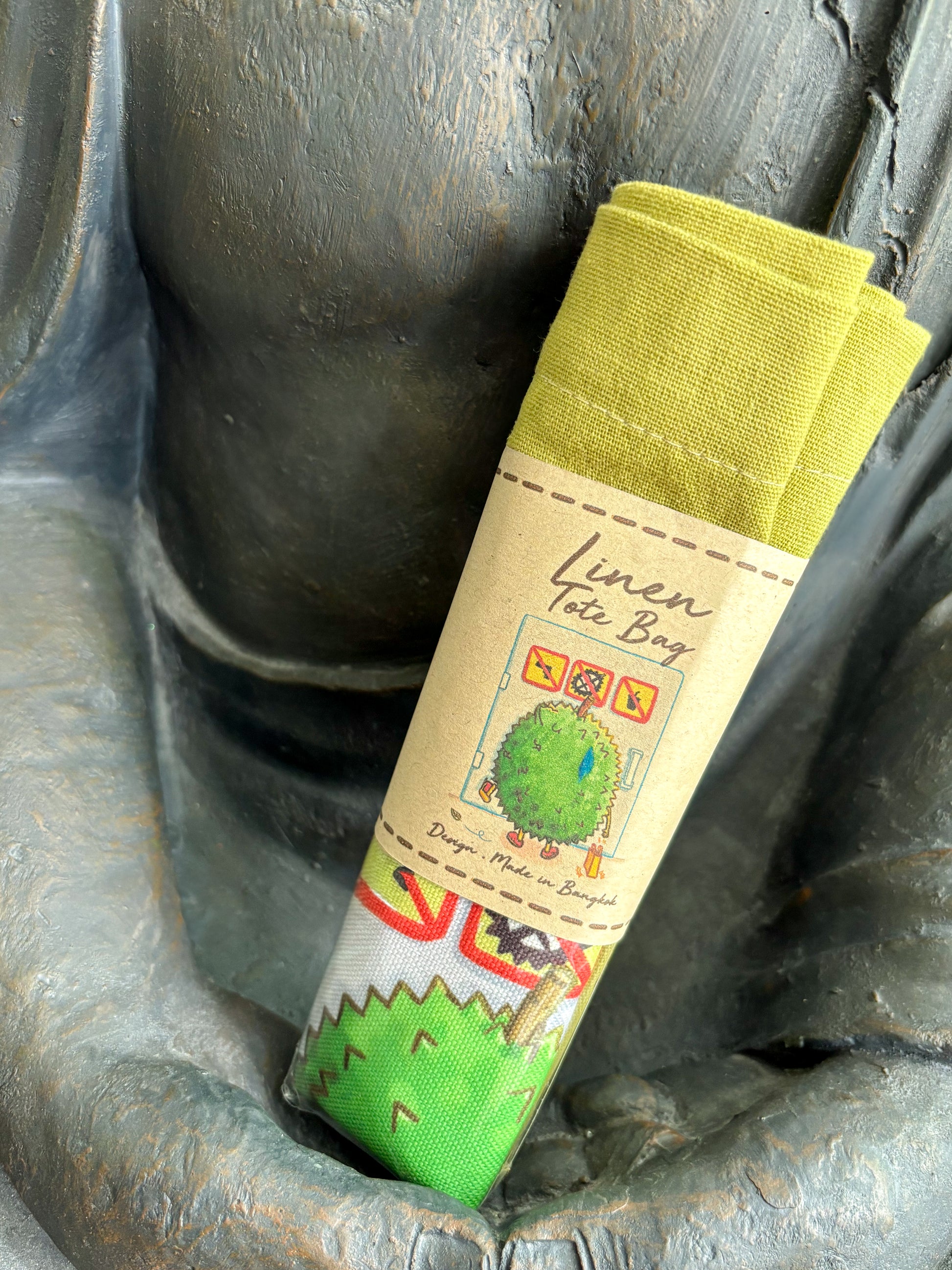 Grüne Leinentasche  zusammengefaltet mit Print von Stinkfrucht Durian die nervös vor Türe steht, wo Verbotsschilder darauf sind, wo Durians verboten sind, Buddhastatue hält das Ganze