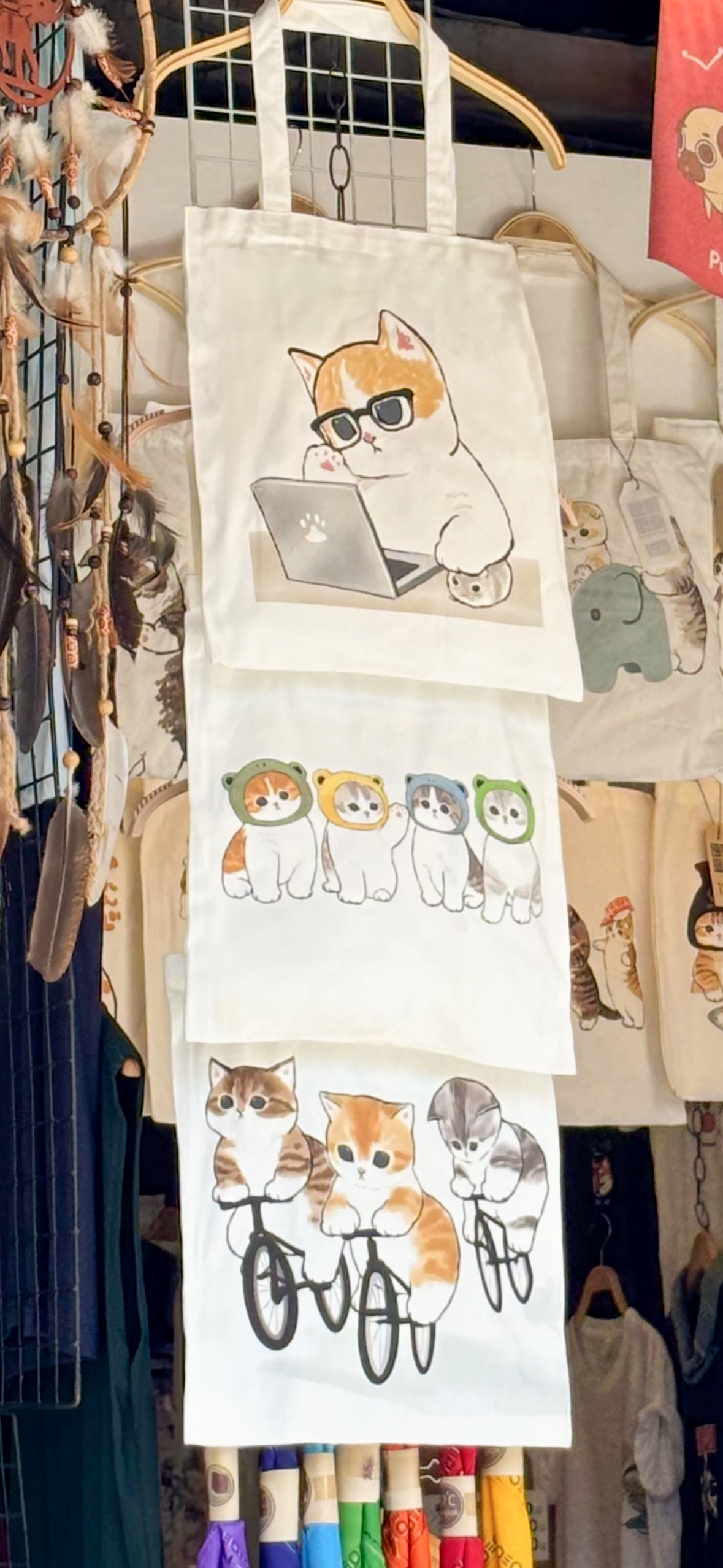Drei aufgehängte weisse Stofftaschen mit lustigen Katzenmotiven – Comic-Katze mit Brille arbeitet am Laptop – perfekte Tote Bag für Katzenliebhaber, IT-Fans und Nerds, zweite Tasche zeigt vier Katzen mit farbigen Hüten und dritte Tasche zeigt drei süsse Katzen auf dem Fahrrad